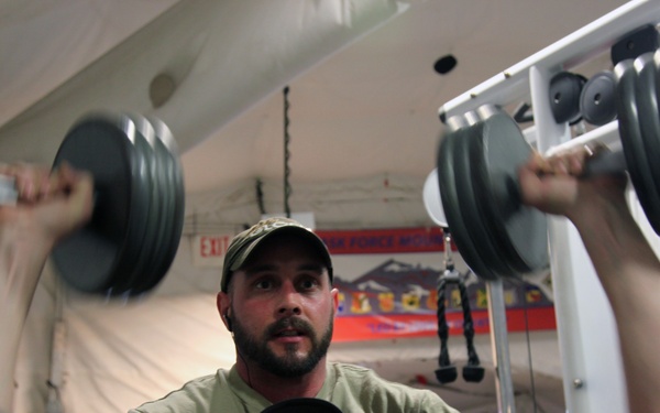 Two men are in a gym setting where one person is lying on a bench performing a dumbbell press exercise while a spotter stands behind him providing assistance. The spotter is wearing a camouflage cap and olive green t-shirt, while the lifter wears a black cap and graphic t-shirt. The gym appears to be located inside a tent or temporary structure, suggesting a military or forward operating base fitness facility.
