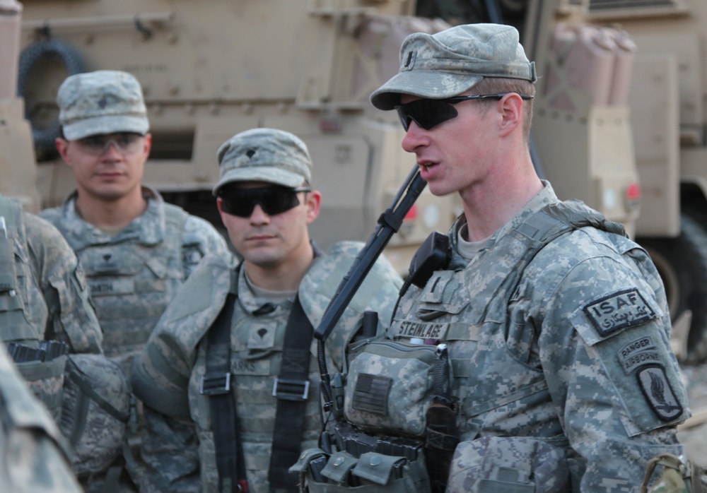 Three U.S. Army soldiers in ACU (Army Combat Uniform) pattern camouflage are gathered in what appears to be a forward operating base or staging area. The soldier in the foreground, identified by his name tape as 'STEINLAGE,' is speaking to the group and wears ISAF, Ranger, and 173rd Airborne Brigade patches. Military vehicles, likely MRAPs or similar armored vehicles, are visible in the background.