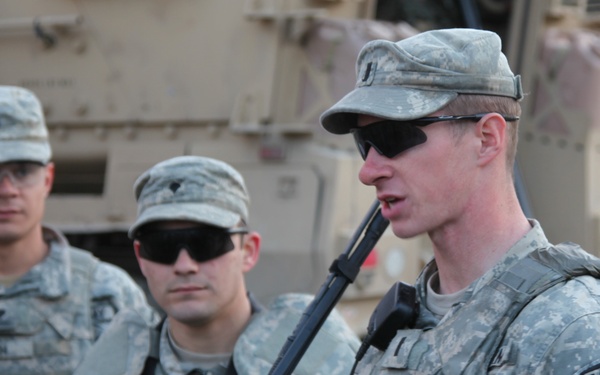 Three U.S. Army soldiers in ACU (Army Combat Uniform) pattern camouflage are gathered in what appears to be a forward operating base or staging area. The soldier in the foreground, identified by his name tape as 'STEINLAGE,' is speaking to the group and wears ISAF, Ranger, and 173rd Airborne Brigade patches. Military vehicles, likely MRAPs or similar armored vehicles, are visible in the background.