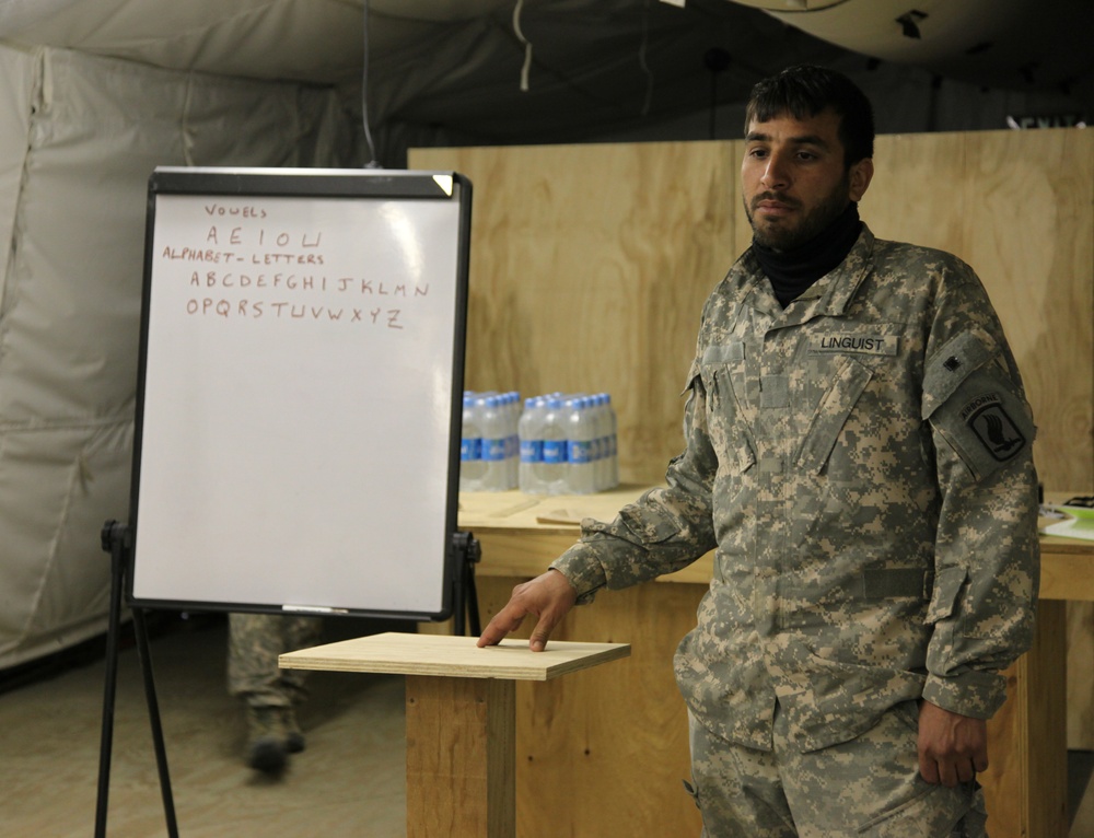 A person wearing US Army ACU (Army Combat Uniform) with a 'LINGUIST' name tape stands next to a whiteboard displaying English vowels and the alphabet. The setting appears to be a forward operating base or military facility with plywood walls and a tent structure. The individual appears to be conducting or preparing for an English language instruction session.