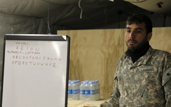 A person wearing US Army ACU (Army Combat Uniform) with a 'LINGUIST' name tape stands next to a whiteboard displaying English vowels and the alphabet. The setting appears to be a forward operating base or military facility with plywood walls and a tent structure. The individual appears to be conducting or preparing for an English language instruction session.