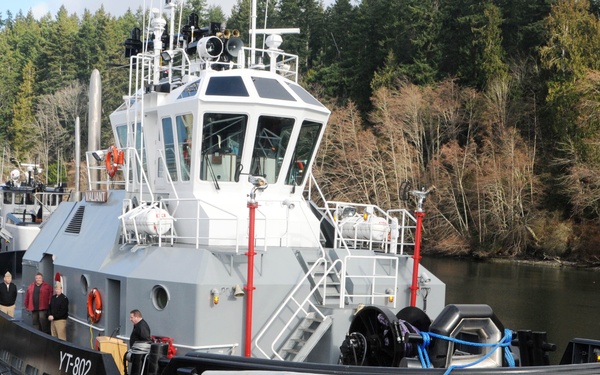 A U.S. Navy harbor tug designated YT-802 is docked at a pier during what appears to be a commissioning or christening ceremony. A crowd of civilians and military personnel are gathered on the dock observing the event. The vessel is a modern gray tugboat with a large rubber fendering system around its hull, red bollards, and a white pilothouse.