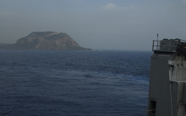 A military naval vessel deck is loaded with multiple tan/desert-colored HMMWVs (Humvees) secured for transport at sea. A vehicle in the foreground bears a red cross marking, indicating a medical/ambulance variant. A rocky island or landmass is visible in the background across open blue ocean water.