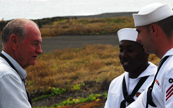 An elderly civilian man in a blue striped shirt is shaking hands with a U.S. Navy sailor in dress white uniform, while a second sailor stands nearby smiling. The interaction appears cordial and takes place outdoors on what looks like a volcanic or coastal terrain with sparse vegetation. The setting suggests a shore visit or commemorative event near a coastline.