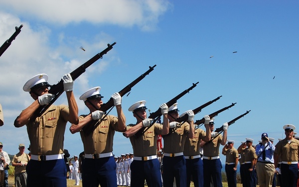A military rifle salute ceremony is being conducted outdoors, likely at a memorial or funeral service. A line of U.S. Marines in dress uniform — tan shirts, blue trousers, and white covers — are firing rifles skyward in a coordinated volley. Spent brass casings are visibly ejecting from the rifles, and additional military personnel and civilians are observing in the background.