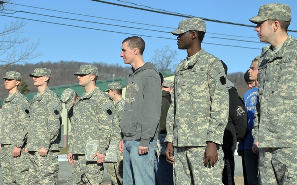 A group of U.S. Army soldiers in ACU (Army Combat Uniform) digital camouflage are standing in formation outdoors during what appears to be a winter or early spring day. Among the uniformed personnel, one individual stands out wearing civilian clothing — a gray hoodie and jeans with white sneakers — suggesting a recruit, visitor, or someone in a non-military role. The setting appears to be a military installation or training area with bare trees, patches of snow, and utility poles visible in the background.