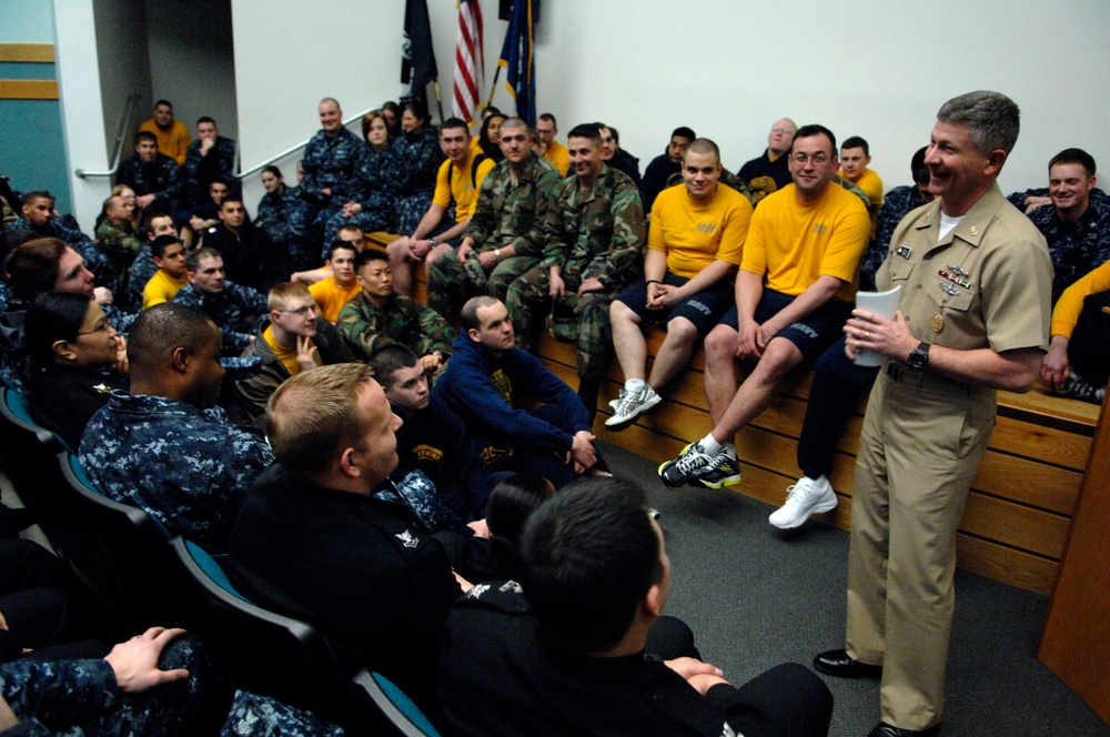 A senior U.S. Navy officer in khaki service uniform addresses a large group of sailors and recruits in an auditorium or briefing room. The audience is dressed in a mix of Navy working uniforms, physical training gear (yellow shirts and shorts), and camouflage uniforms. The atmosphere appears informal and engaging, with visible smiling and laughter among attendees.