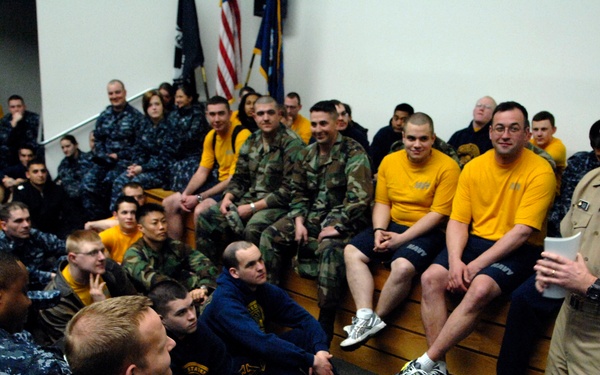 A senior U.S. Navy officer in khaki service uniform addresses a large group of sailors and recruits in an auditorium or briefing room. The audience is dressed in a mix of Navy working uniforms, physical training gear (yellow shirts and shorts), and camouflage uniforms. The atmosphere appears informal and engaging, with visible smiling and laughter among attendees.