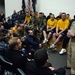 A senior U.S. Navy officer in khaki service uniform addresses a large group of sailors and recruits in an auditorium or briefing room. The audience is dressed in a mix of Navy working uniforms, physical training gear (yellow shirts and shorts), and camouflage uniforms. The atmosphere appears informal and engaging, with visible smiling and laughter among attendees.