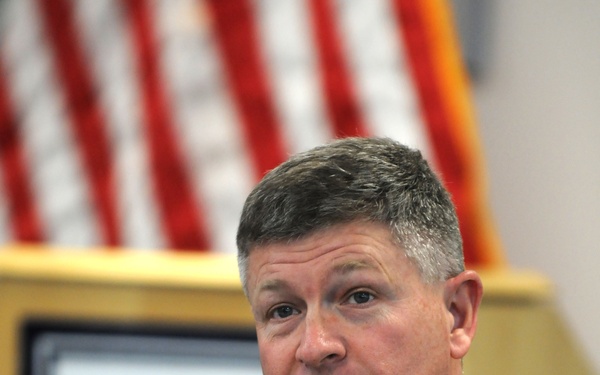 A U.S. Navy officer in khaki service uniform is seated and speaking, gesturing with one hand while holding a pen in the other. He is in what appears to be a briefing room or conference setting with an American flag and a military unit insignia plaque visible in the background. His uniform displays several ribbons, warfare qualification badges, and rank insignia.
