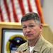 A U.S. Navy officer in khaki service uniform is seated and speaking, gesturing with one hand while holding a pen in the other. He is in what appears to be a briefing room or conference setting with an American flag and a military unit insignia plaque visible in the background. His uniform displays several ribbons, warfare qualification badges, and rank insignia.