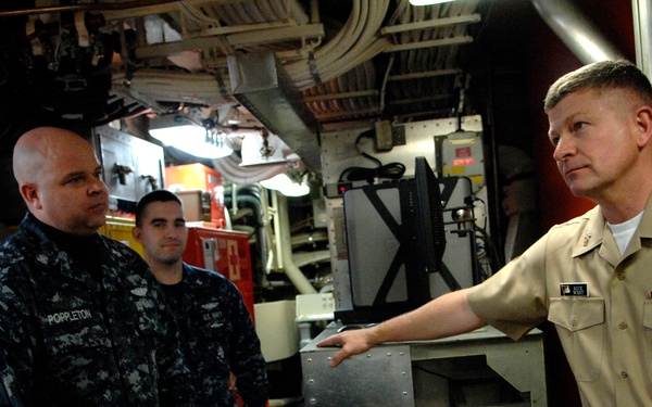 A senior U.S. Navy officer in khaki service uniform is briefing or instructing several enlisted sailors in Navy Working Uniform (NWU) camouflage inside what appears to be a shipboard compartment or engine room. The officer is gesturing toward equipment while the sailors listen attentively. The setting is a confined, industrial naval vessel interior with visible safety and mechanical equipment.