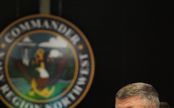 A military officer in a dark uniform sits attentively at a conference table with hands clasped near his chin in a thoughtful pose. Behind him is the official seal of 'Commander, Navy Region Northwest' displayed on a dark backdrop. Other personnel are visible in the background, and binders/documents are present on the table in the foreground.