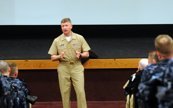A senior U.S. Navy officer in khaki service uniform is addressing a large group of sailors seated in rows within an auditorium or briefing hall. The speaker stands at the front between two columns of seated personnel wearing Navy Working Uniform (NWU) digital blue camouflage. The setting appears to be a formal military briefing or all-hands meeting.