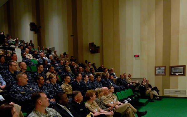 A military briefing or all-hands meeting is taking place in a formal auditorium setting. A senior officer in tan/khaki service dress uniform stands at the front addressing a large audience of military personnel seated in green theater-style chairs. The audience includes members from multiple branches, predominantly wearing Navy working uniforms (NWU) with some Army combat uniforms visible.