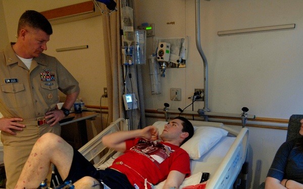 A military officer in U.S. Navy service uniform visits a young male patient in a hospital room. The patient, wearing a red shirt, is lying in a hospital bed with visible wounds and medical devices on his legs, while a young woman sits nearby in a chair. The scene appears to be a bedside visit, likely at a military medical facility.