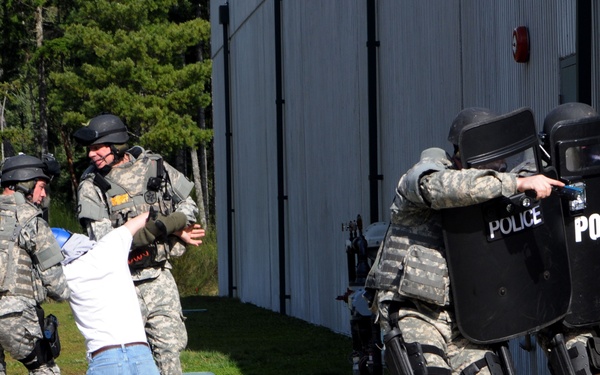 The image depicts what appears to be a law enforcement or military training exercise. Personnel in camouflage uniforms and tactical gear, including ballistic shields marked 'POLICE,' are detaining a civilian role-player who is on their knees. The setting is outdoors near a metal building with a treeline in the background.