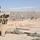 A military construction site in an arid desert environment, likely a forward operating base. Personnel in military camouflage uniforms are working with lumber and building materials within a perimeter secured by HESCO barriers and fencing. A handwritten sign in the foreground warns of the construction area and hard hat requirements.