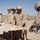 A group of U.S. Marines in desert camouflage uniforms and combat helmets are engaged in a carpentry or construction task at what appears to be a forward operating base in an arid environment. One Marine on the right is operating a DeWalt circular saw to cut lumber while others hold the wood steady on a makeshift workbench. The background shows a perimeter wall, flags, and additional personnel working in the area.