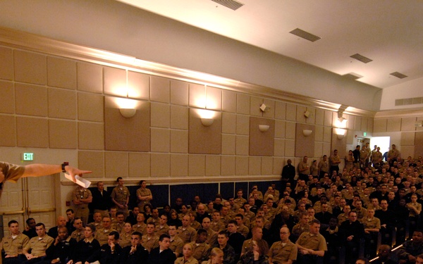 A military officer in tan/khaki uniform stands on a raised wooden stage addressing a large audience of uniformed military personnel in an auditorium or assembly hall. The speaker is holding a microphone and gesturing toward the seated crowd, which fills the room to capacity. The setting appears to be a formal military all-hands briefing or command presentation.