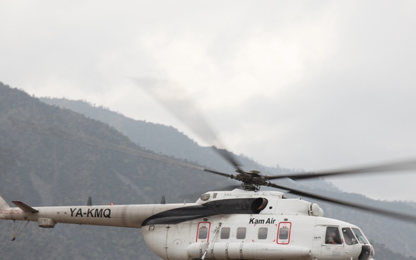 A white Mil Mi-8 helicopter operated by Kam Air, registered YA-KMQ, is captured in low-altitude flight against a mountainous backdrop. The helicopter appears to be either landing or taking off in a rugged, hilly terrain environment. The overcast sky and misty mountains suggest a high-altitude location, likely in Afghanistan given the Afghan registration prefix 'YA'.