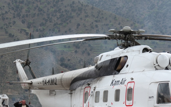A white Kam Air helicopter with registration YA-KMQ is parked on a concrete pad in a mountainous region. Two individuals stand near the aircraft alongside a Bobcat S330 skid-steer loader. The setting appears to be a remote helipad or small airstrip surrounded by rugged, sparsely vegetated hills.