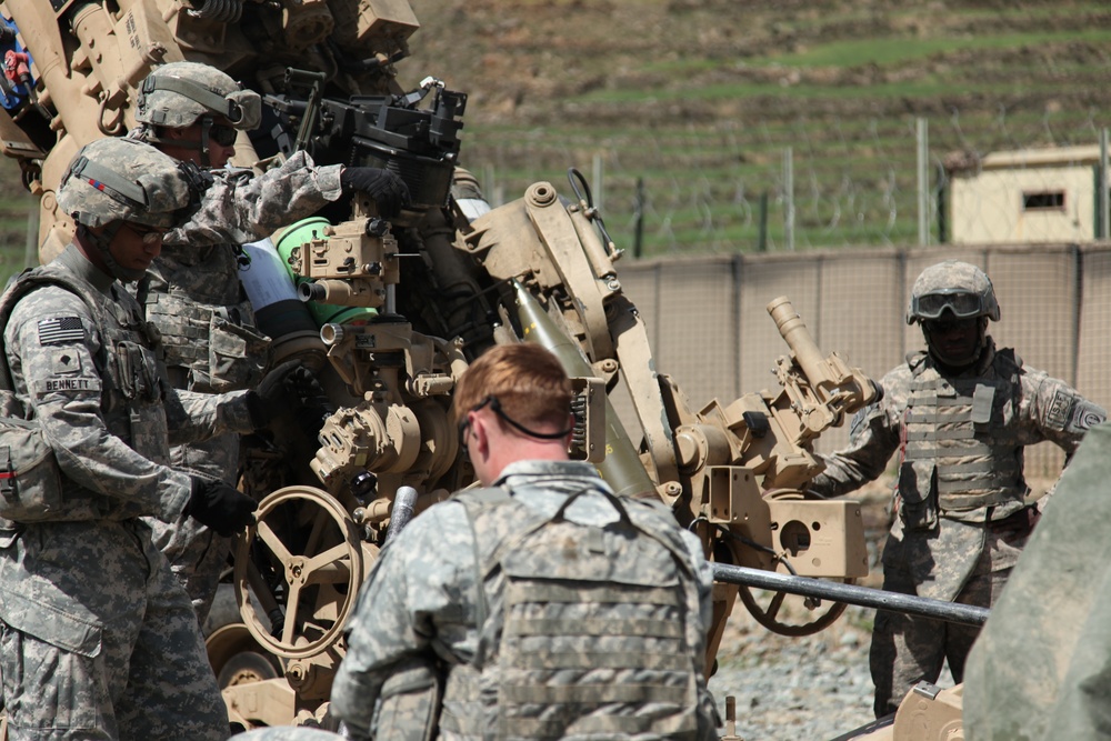 U.S. Army soldiers in ACU (Army Combat Uniform) are working around what appears to be an M777 howitzer or similar artillery piece in a forward operating base or training environment. The soldiers are engaged in what appears to be artillery emplacement, maintenance, or training operations. A guard tower and fenced perimeter are visible in the background, suggesting a military installation.