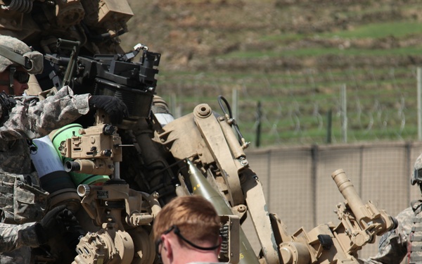 U.S. Army soldiers in ACU (Army Combat Uniform) are working around what appears to be an M777 howitzer or similar artillery piece in a forward operating base or training environment. The soldiers are engaged in what appears to be artillery emplacement, maintenance, or training operations. A guard tower and fenced perimeter are visible in the background, suggesting a military installation.
