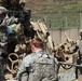 U.S. Army soldiers in ACU (Army Combat Uniform) are working around what appears to be an M777 howitzer or similar artillery piece in a forward operating base or training environment. The soldiers are engaged in what appears to be artillery emplacement, maintenance, or training operations. A guard tower and fenced perimeter are visible in the background, suggesting a military installation.