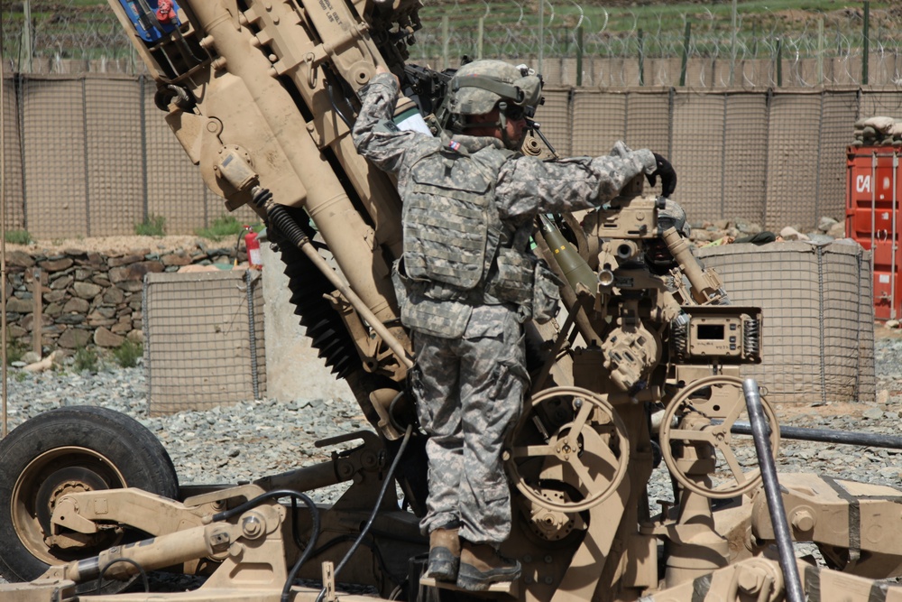 A U.S. Army soldier in ACU (Army Combat Uniform) pattern camouflage is working on what appears to be an M777 lightweight howitzer or similar artillery piece at a forward operating base. The soldier is wearing full combat gear including a helmet, body armor, and tactical vest. The setting appears to be a fortified military installation, likely in Afghanistan based on the terrain and infrastructure visible.