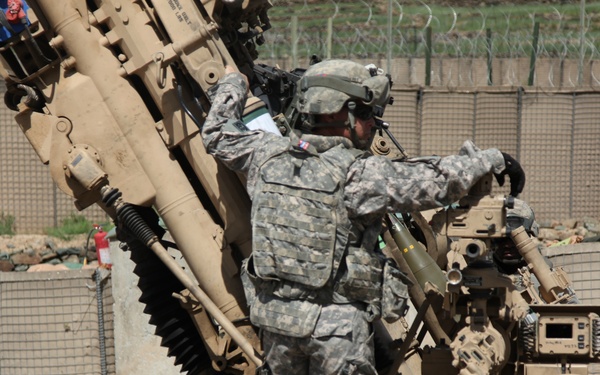 A U.S. Army soldier in ACU (Army Combat Uniform) pattern camouflage is working on what appears to be an M777 lightweight howitzer or similar artillery piece at a forward operating base. The soldier is wearing full combat gear including a helmet, body armor, and tactical vest. The setting appears to be a fortified military installation, likely in Afghanistan based on the terrain and infrastructure visible.