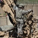 A U.S. Army soldier in ACU (Army Combat Uniform) pattern camouflage is working on what appears to be an M777 lightweight howitzer or similar artillery piece at a forward operating base. The soldier is wearing full combat gear including a helmet, body armor, and tactical vest. The setting appears to be a fortified military installation, likely in Afghanistan based on the terrain and infrastructure visible.