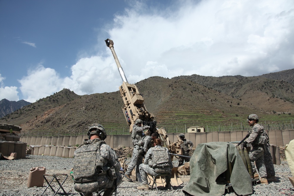The image shows U.S. Army soldiers operating what appears to be an M777 155mm howitzer at a forward operating base in a mountainous region, consistent with Afghanistan. The artillery piece is elevated at a high angle, suggesting it is either being prepared for firing or has recently been fired. Several soldiers in ACU (Army Combat Uniform) pattern camouflage are positioned around the weapon system within a fortified compound.