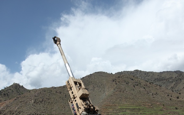 The image shows U.S. Army soldiers operating what appears to be an M777 155mm howitzer at a forward operating base in a mountainous region, consistent with Afghanistan. The artillery piece is elevated at a high angle, suggesting it is either being prepared for firing or has recently been fired. Several soldiers in ACU (Army Combat Uniform) pattern camouflage are positioned around the weapon system within a fortified compound.