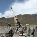 The image shows U.S. Army soldiers operating what appears to be an M777 155mm howitzer at a forward operating base in a mountainous region, consistent with Afghanistan. The artillery piece is elevated at a high angle, suggesting it is either being prepared for firing or has recently been fired. Several soldiers in ACU (Army Combat Uniform) pattern camouflage are positioned around the weapon system within a fortified compound.