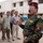 Alaskan MPs run Iraqi Police through tough training A military officer in camouflage uniform and red beret is addressing a group of individuals in an outdoor compound setting. The central figure wears Iraqi Special Forces patches and insignia, and appears to be briefing or speaking to a mixed group of uniformed and civilian-dressed personnel. Military vehicles are visible in the background near a multi-story building.