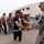 Alaskan MPs run Iraqi Police through tough training A U.S. Army Military Police (MP) soldier in ACU camouflage uniform is shaking hands with a civilian man in a black tracksuit, while a group of civilians stands in a queue behind him. The setting appears to be an outdoor area near a large concrete building, likely in a Middle Eastern location based on the architecture and environment. The scene suggests a processing, registration, or community engagement event.