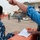 Alaskan MPs run Iraqi Police through tough training The image shows a group of individuals in a military or security compound environment, with some wearing blue camouflage uniforms and others in civilian clothing. A person in a blue shirt is pointing and appears to be directing or instructing others while referencing a document on an orange barrier. The background shows a gravel compound with military tents, concertina wire, concrete barriers, and personnel working in the area.