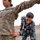 Alaskan MPs run Iraqi Police through tough training A U.S. Army soldier (female, wearing ACU digital camouflage) is being searched or pat-down trained by a male officer wearing Iraqi Ministry of Interior police uniform. The scene appears to be a training exercise at a military or police compound, with additional personnel observing in the background. The setting includes concrete barriers, razor wire, and a gravel surface consistent with a forward operating base or training facility.