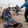 Alaskan MPs run Iraqi Police through tough training The image shows what appears to be a military or police training exercise at a secured compound with razor wire fencing. Personnel in blue camouflage uniforms, with visible 'POLICE' patches and unit insignia, are practicing casualty response or detainee handling procedures. Multiple individuals are involved in the drill, including one person on the ground, one kneeling beside them, and others directing or observing.