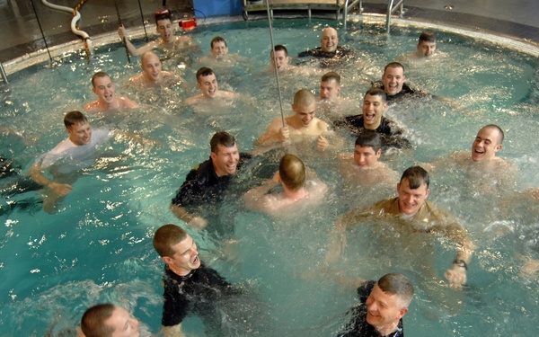 A large group of approximately 30-35 men are gathered in a circular indoor pool, many appearing to be laughing and celebrating. Some individuals are wearing black clothing while others appear shirtless, suggesting a mix of instructors and participants. The atmosphere appears festive and energetic, consistent with a graduation or completion ceremony.