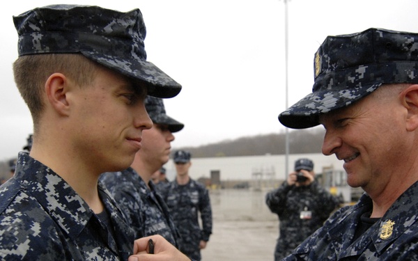 A senior U.S. Navy enlisted or officer personnel is pinning a medal or device onto the uniform of a younger sailor named 'CLIBER' during what appears to be a formal military ceremony. The event is taking place outdoors at what appears to be a naval installation or pier, with a formation of sailors visible in the background. A photographer is documenting the ceremony.