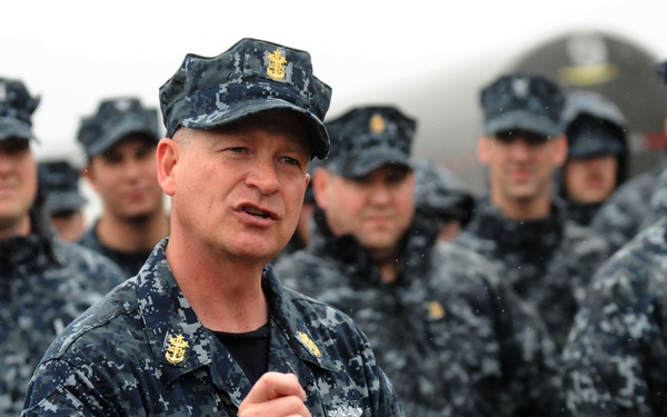 A senior U.S. Navy enlisted leader, identifiable by gold anchors on his collar and cap insignia consistent with a Master Chief Petty Officer, appears to be addressing a formation of sailors. The personnel are all wearing the Navy Working Uniform (NWU) Type I, the blue digital camouflage pattern. The setting appears to be an outdoor naval facility, possibly during a muster or briefing.