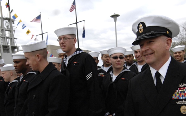 A group of U.S. Navy personnel are assembled in formation, wearing dress blue uniforms with white Dixie cup covers. A senior enlisted officer, identifiable by his combination cover, multiple service ribbons, and command insignia, stands prominently in the foreground. The setting appears to be a naval installation or pier area with signal flags and a ship's superstructure visible in the background.