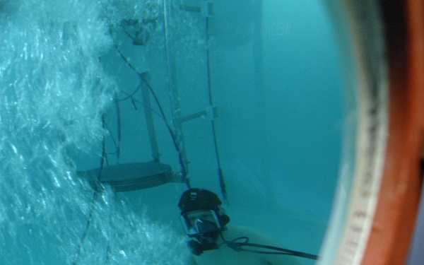 The image is taken through a circular porthole window, showing an underwater view of a person submerged in what appears to be a training or research pool. The individual is wearing a diving mask and dark shorts, surrounded by rising air bubbles. The facility appears to be an indoor aquatic training tank, likely used for military, scientific, or emergency response training.