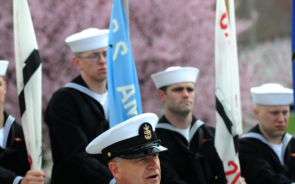A senior U.S. Navy officer, wearing a dress uniform with multiple service ribbons and a chief petty officer or commissioned officer cover, speaks at a podium during a formal outdoor ceremony. Behind him, a color guard of enlisted sailors in dress blue uniforms hold various flags, including what appears to be unit or ship pennants. Cherry blossom trees are visible in the background, suggesting a spring setting.
