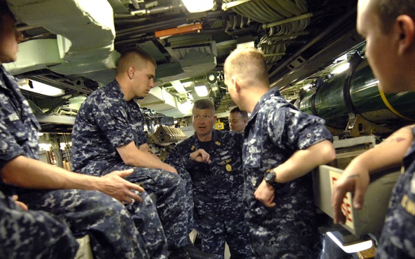 A group of U.S. Navy personnel in Navy Working Uniform (NWU) Type I blue digital camouflage are gathered in a confined interior space, consistent with a submarine torpedo room. A senior enlisted or officer figure appears to be briefing or instructing the group, with large green cylindrical objects (likely torpedoes or torpedo tubes) visible in the background. The cramped, equipment-dense environment is characteristic of a submarine's forward compartment.