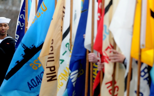 A U.S. Navy sailor in dress uniform stands at attention among a large display of military unit and organizational flags at what appears to be a memorial or ceremonial event. The flags represent various naval vessels and units, including a visible flag for 'USS SS-275 New York (East)'. Small American flags are also visible in the background near what appears to be a memorial or grave site.