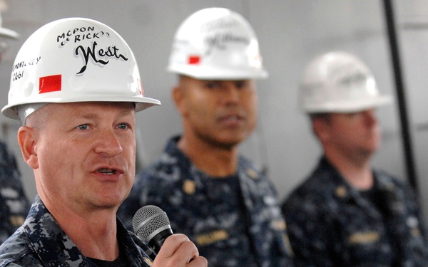 A U.S. Navy senior enlisted leader wearing a white hard hat labeled 'MCPON Rick West' is speaking into a microphone while addressing a group of sailors. The speaker is in the foreground wearing Navy Working Uniform (NWU) digital camouflage and appears to be giving a speech or briefing. Several other sailors in identical uniforms and white hard hats stand attentively in the background.