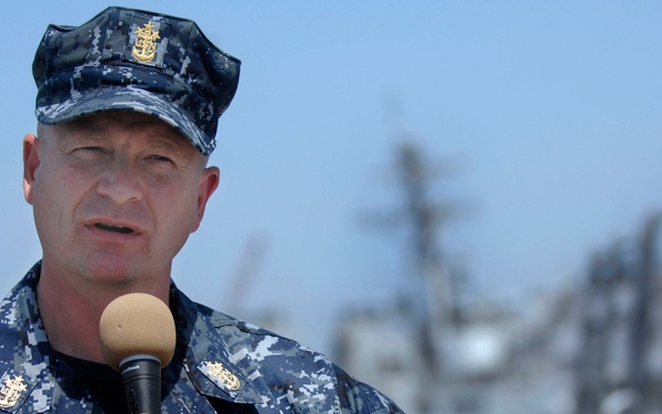 A U.S. Navy senior enlisted personnel is speaking into a microphone, likely addressing an audience at a naval facility or aboard a ship. The individual is wearing the Navy Working Uniform (NWU) digital camouflage pattern with visible rank insignia and qualification badges. Naval vessels are visible in the blurred background, suggesting a pier or shipyard setting.